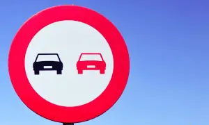 Señal de tráfico: dos coches, uno negro y otro rojo, en un círculo rojo sobre fondo blanco. Indica donde está prohibido adelantar.