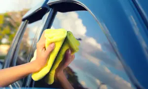 Manos limpiando los cristales del coche con un paño amarillo. El cielo azul y las nubes se reflejan en el cristal.