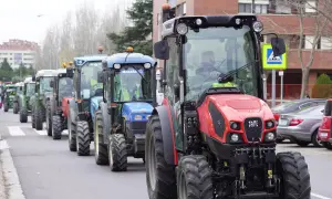 Una fila de tractores, algunos verdes y uno rojo al frente, circulan por una calle urbana. Parece que un tractor puede ir por autovía.