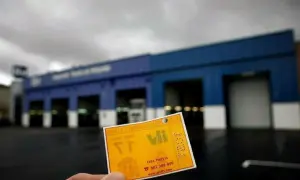 Tarjeta amarilla de cita previa para la ITV, con el logo de V.I. y el número de teléfono. Al fondo, el edificio de la estación de inspección técnica de vehículos.