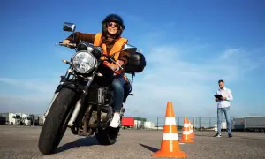 Mujer sonriente con casco y chaleco naranja en moto, lista para obtener el carnet A. Instructor observa.