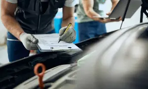 Mecánico con guantes revisa papeles remolque mientras colega usa portátil.