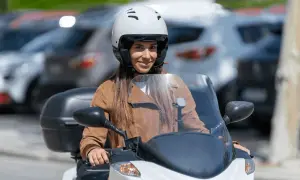 Mujer sonriente con casco blanco y chaqueta marrón en moto. Averigua el precio del carnet de moto A2.