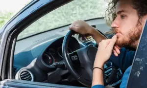 Hombre con barba y pelo rizado, pensativo, con la mano en la barbilla, dentro de un coche. Se escucha un ruido clack clack al girar el volante en parado.
