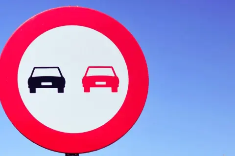 Señal de tráfico: dos coches, uno negro y otro rojo, en un círculo rojo sobre fondo blanco. Indica donde está prohibido adelantar.