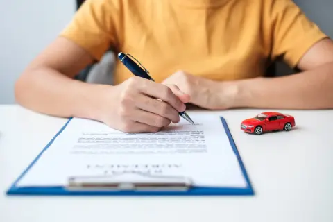 Persona firma un documento, quizás para saber como dar de baja un seguro de coche. Un coche rojo en miniatura está al lado.