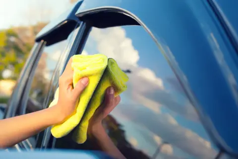 Manos limpiando los cristales del coche con un paño amarillo. El cielo azul y las nubes se reflejan en el cristal.