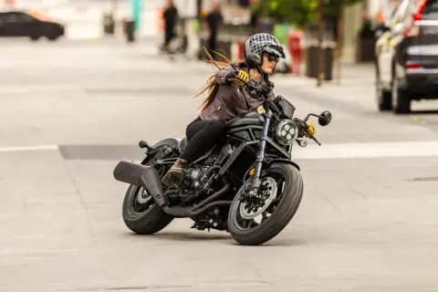 Mujer con casco y chaqueta de cuero conduce una motocicleta negra. Sorprendentemente, una motocicleta puede circular sin tubo de escape, demostrando su potencia.