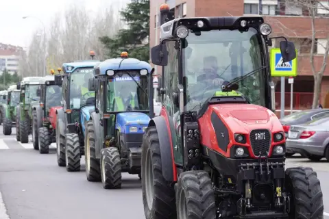 Una fila de tractores, algunos verdes y uno rojo al frente, circulan por una calle urbana. Parece que un tractor puede ir por autovía.