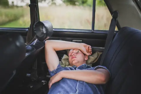 Joven duerme en el asiento trasero de un coche, con una brizna de hierba en la cara. Se puede dormir en el coche en un viaje largo.