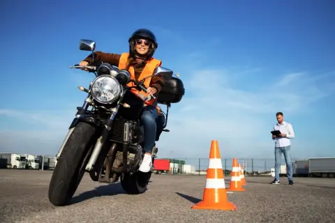 Mujer sonriente con casco y chaleco naranja en moto, lista para obtener el carnet A. Instructor observa.