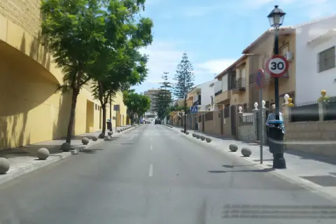 Calle soleada en zbe Fuengirola con casas blancas y amarillas, árboles verdes y bolardos de hormigón.