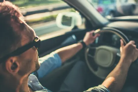 Hombre con gafas de sol conduce, notando que mi coche vibra a partir de 100 km/h. El sol ilumina su cabello.
