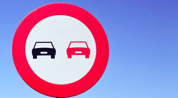 Señal de tráfico: dos coches, uno negro y otro rojo, en un círculo rojo sobre fondo blanco. Indica donde está prohibido adelantar.