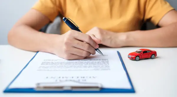 Persona firma un documento, quizás para saber como dar de baja un seguro de coche. Un coche rojo en miniatura está al lado.