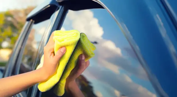 Manos limpiando los cristales del coche con un paño amarillo. El cielo azul y las nubes se reflejan en el cristal.