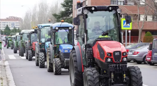 Una fila de tractores, algunos verdes y uno rojo al frente, circulan por una calle urbana. Parece que un tractor puede ir por autovía.