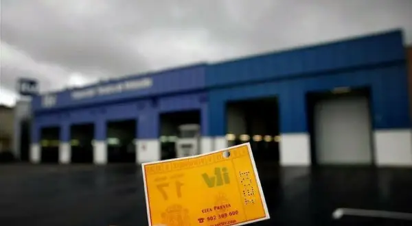 Tarjeta amarilla de cita previa para la ITV, con el logo de V.I. y el número de teléfono. Al fondo, el edificio de la estación de inspección técnica de vehículos.