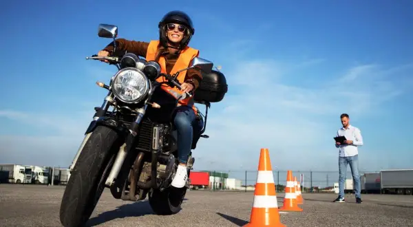 Mujer sonriente con casco y chaleco naranja en moto, lista para obtener el carnet A. Instructor observa.