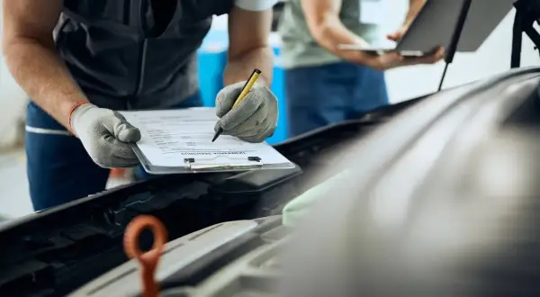 Mecánico con guantes revisa papeles remolque mientras colega usa portátil.