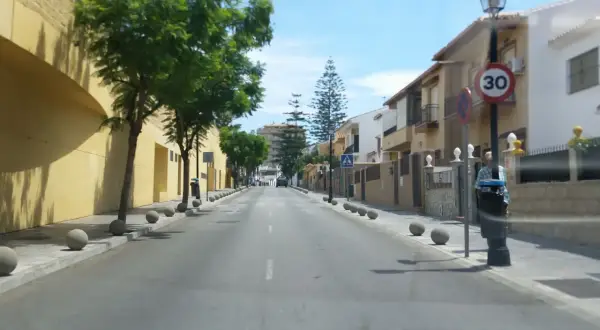 Calle soleada en zbe Fuengirola con casas blancas y amarillas, árboles verdes y bolardos de hormigón.