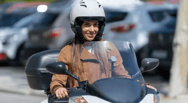 Mujer sonriente con casco blanco y chaqueta marrón en moto. Averigua el precio del carnet de moto A2.