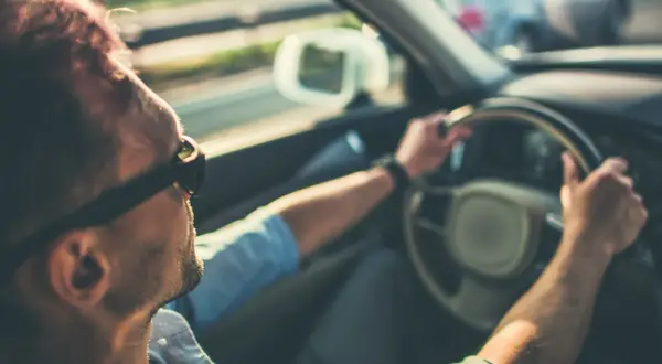 Hombre con gafas de sol conduce, notando que mi coche vibra a partir de 100 km/h. El sol ilumina su cabello.