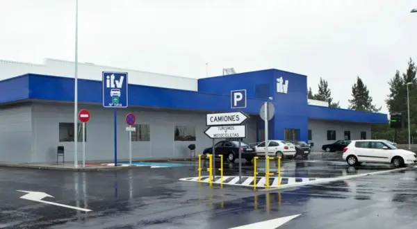 Edificio azul y gris de **itv extraordinaria** con señales de tráfico y coches aparcados en un día lluvioso.