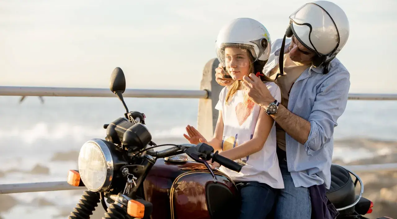 Un padre ayuda a su hija a ponerse el casco para un paseo en moto. Ambos llevan cascos blancos.