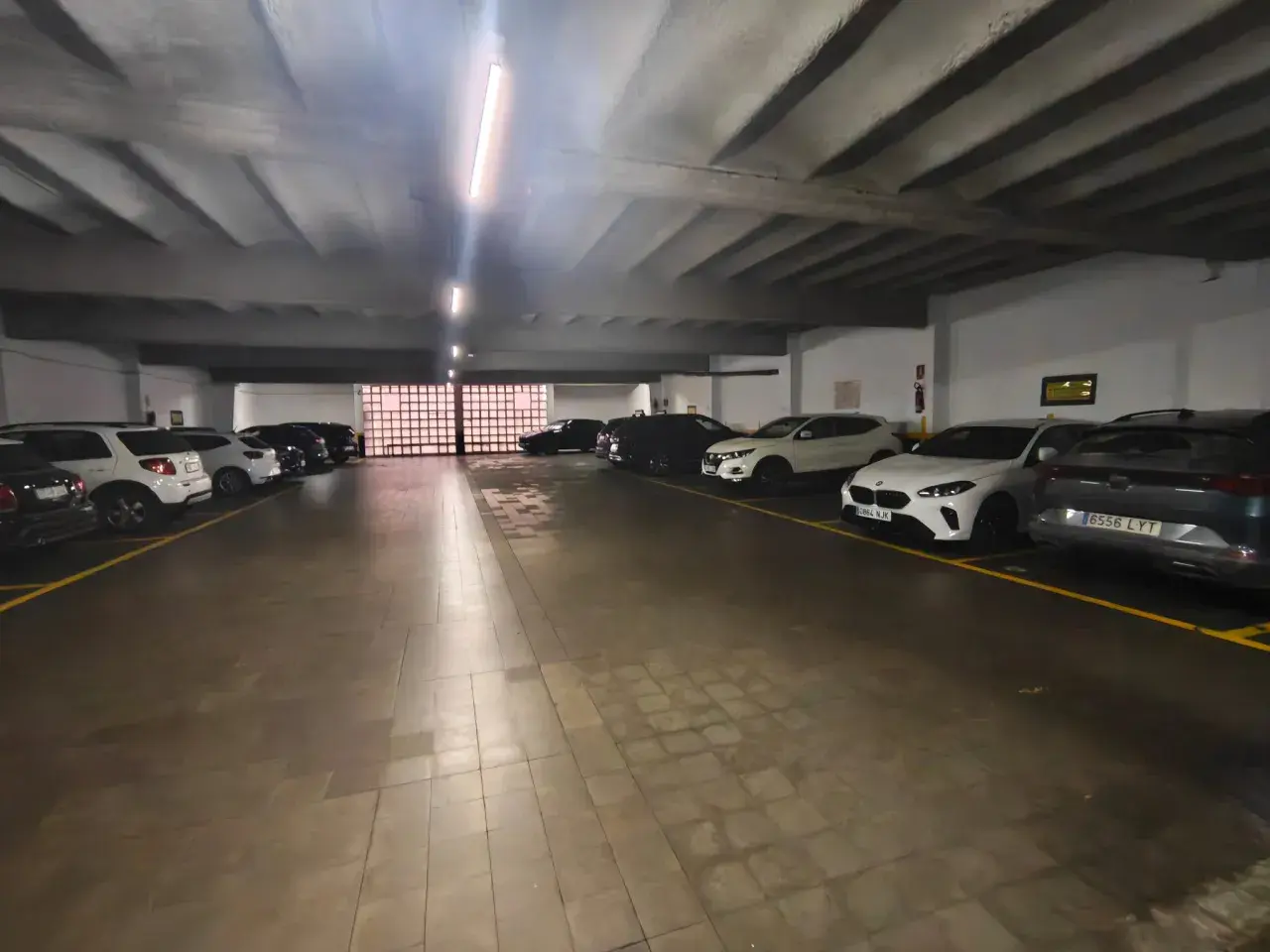 Coches aparcados en diagonal en un garaje subterráneo con iluminación fluorescente y suelo de baldosas.