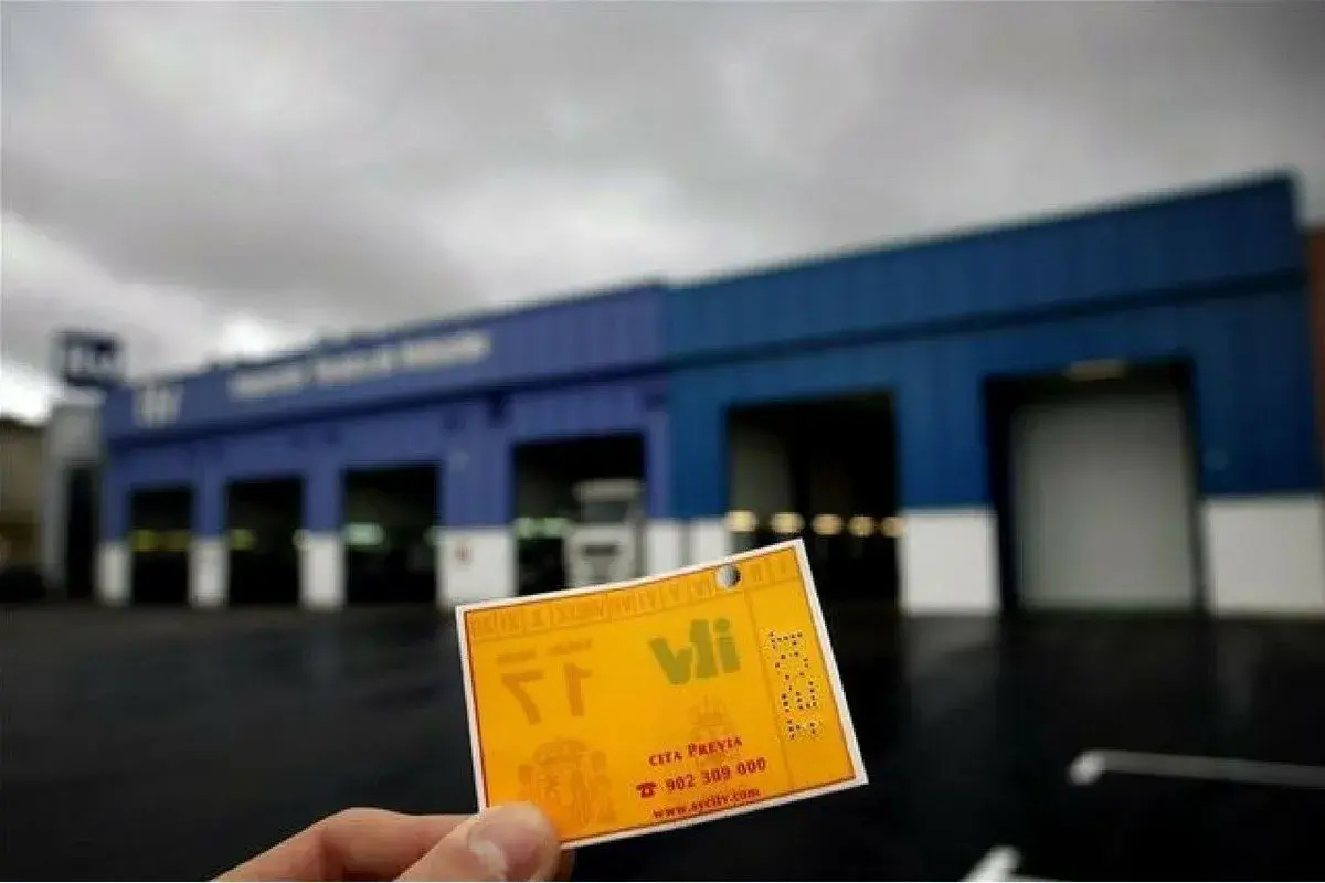 Tarjeta amarilla de cita previa para la ITV, con el logo de V.I. y el número de teléfono. Al fondo, el edificio de la estación de inspección técnica de vehículos.