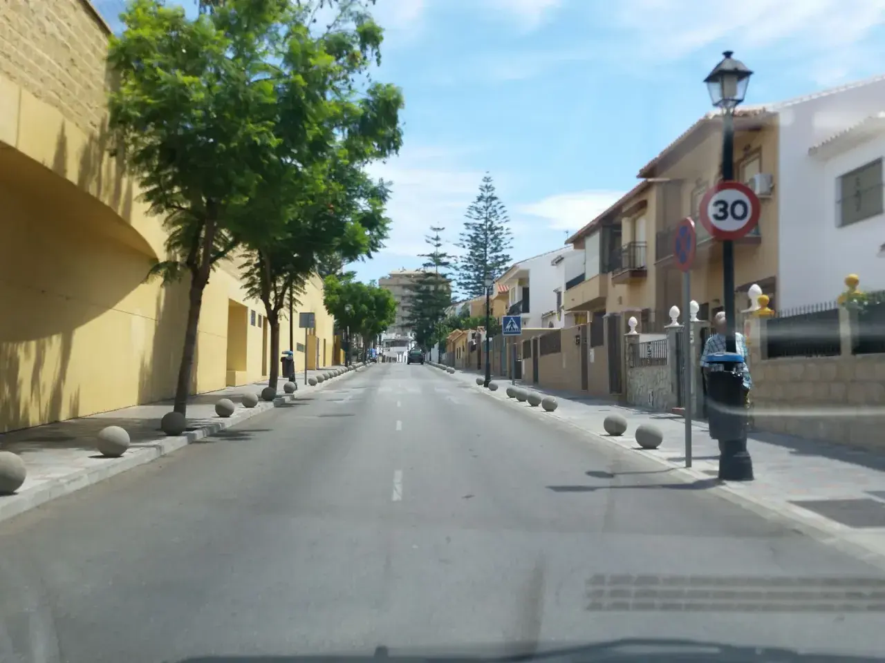 Calle soleada en zbe Fuengirola con casas blancas y amarillas, árboles verdes y bolardos de hormigón.