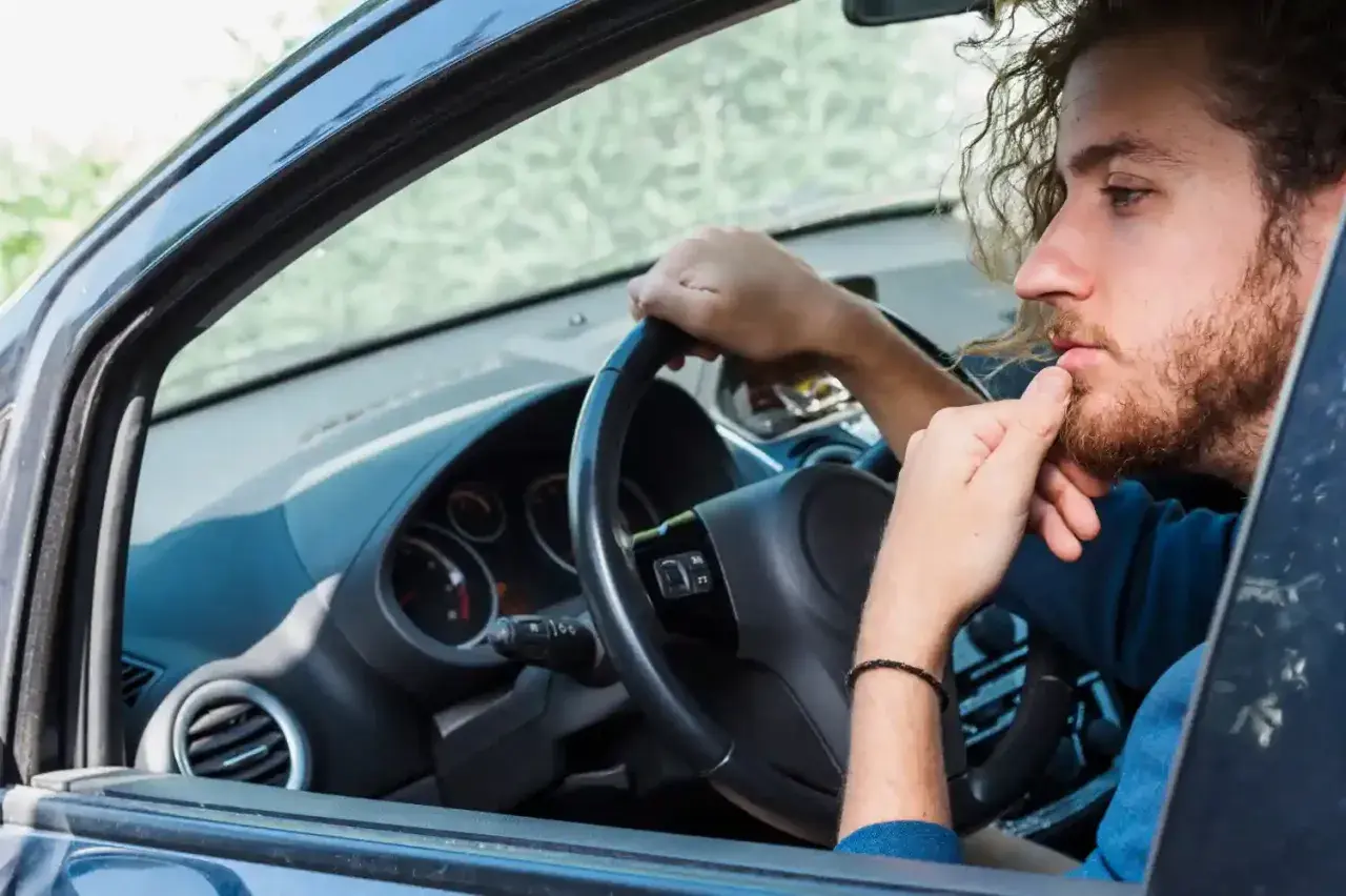 Hombre con barba y pelo rizado, pensativo, con la mano en la barbilla, dentro de un coche. Se escucha un ruido clack clack al girar el volante en parado.