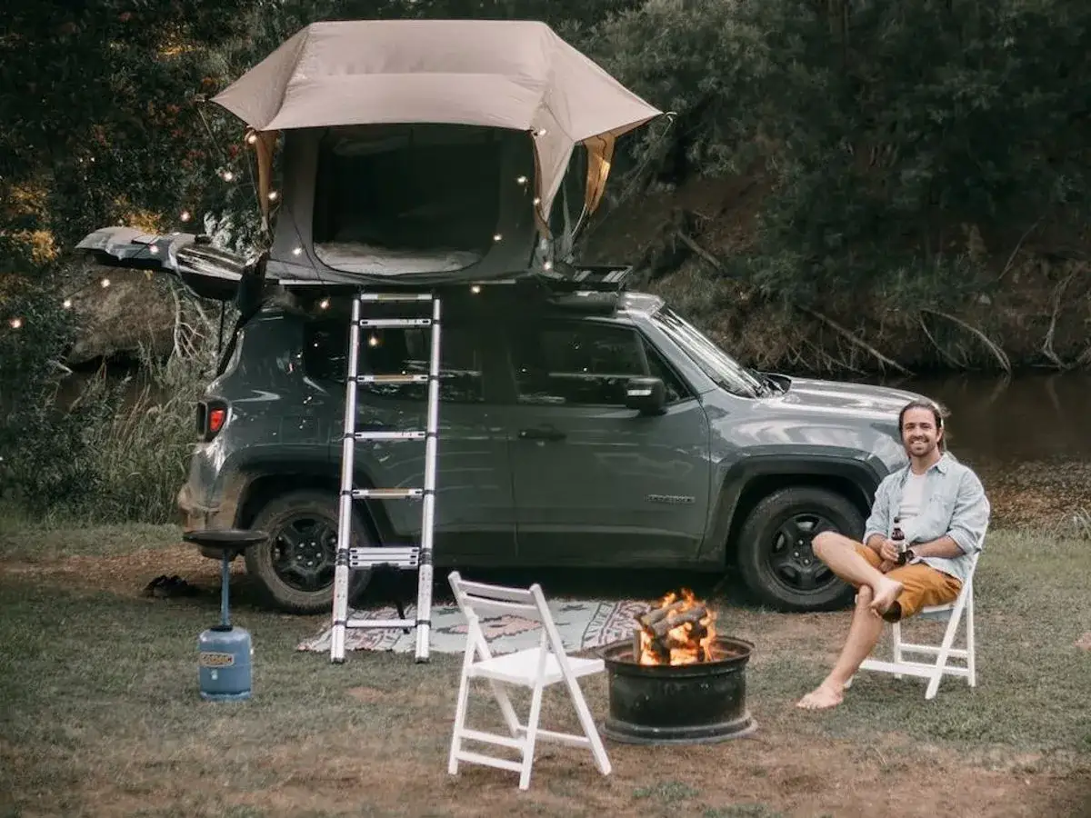 Hombre sonr&iacute;e junto a fogata, con tienda de techo en coche. &iexcl;Perfecto para cuando se puede dormir en el coche!