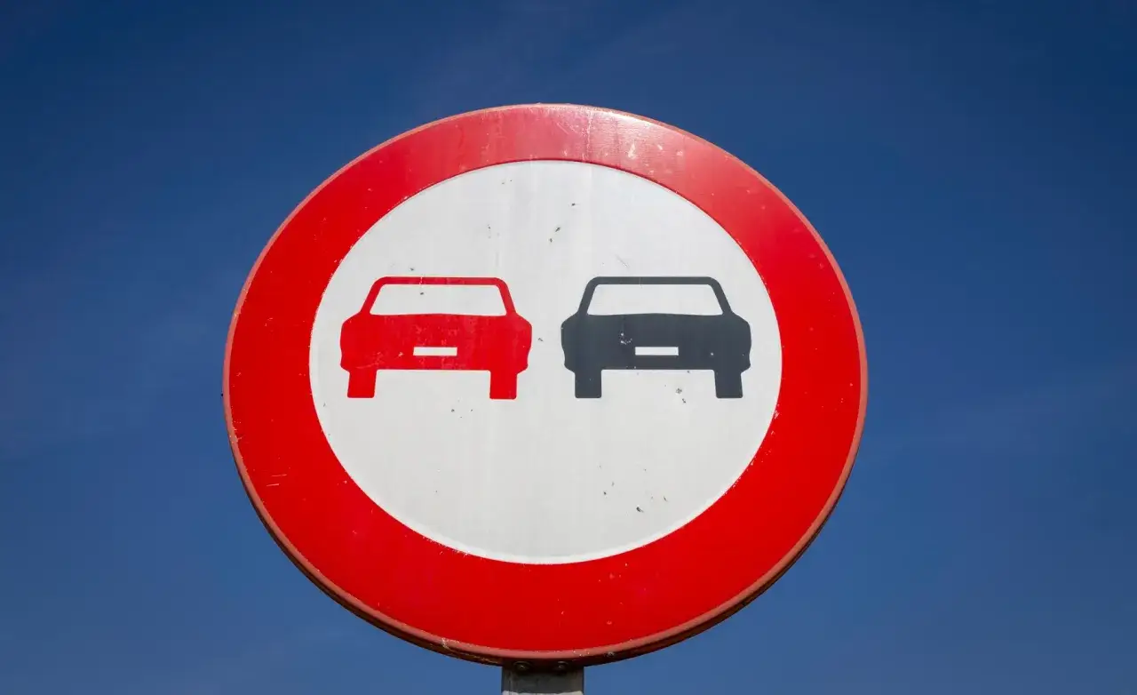Señal de tráfico circular con borde rojo y fondo blanco, que muestra dos coches uno al lado del otro. Indica una zona donde está prohibido adelantar.