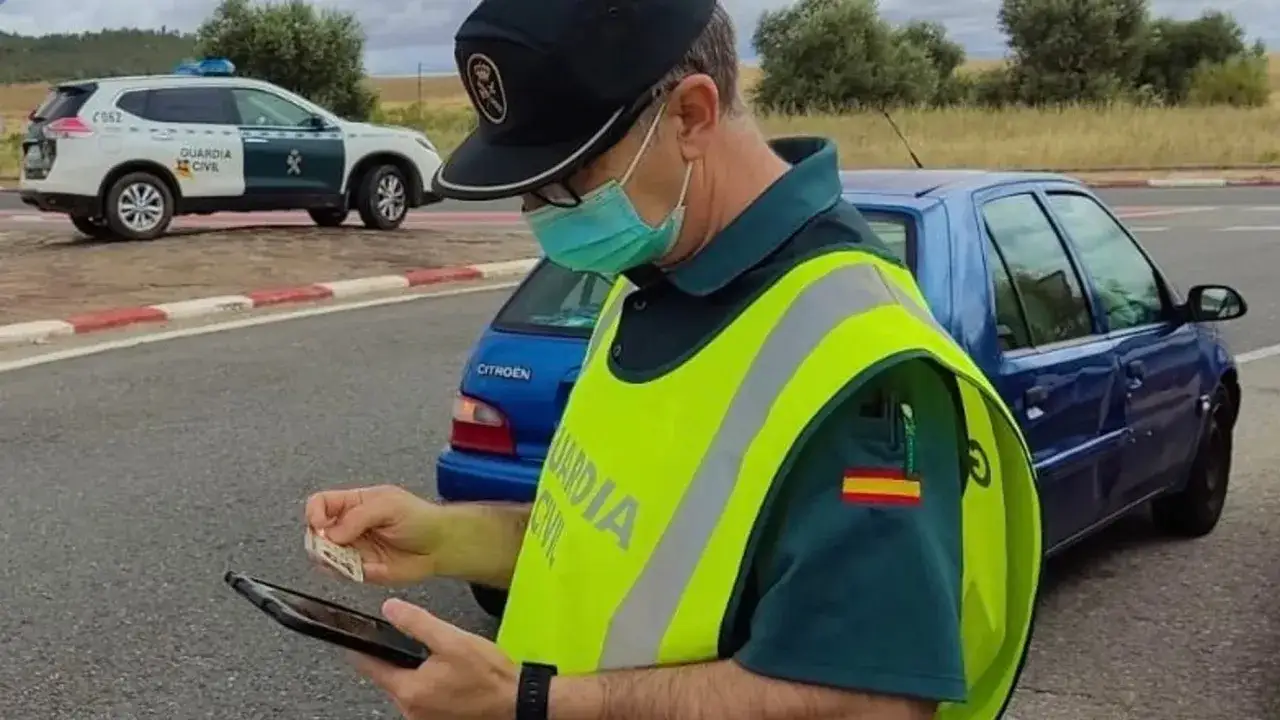 Agente de la Guardia Civil con mascarilla revisa documentación en su tablet. A su lado, un coche azul y un vehículo oficial de la Guardia Civil. Fiva comprobar seguro.