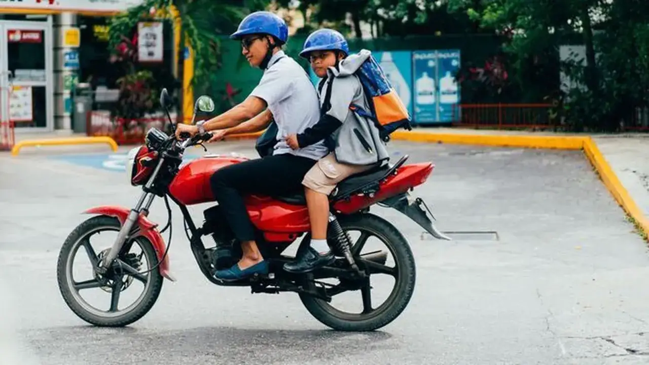 Un hombre y un niño en un ciclomotor rojo, ambos con cascos azules, circulan por una calle. El niño lleva una mochila.
