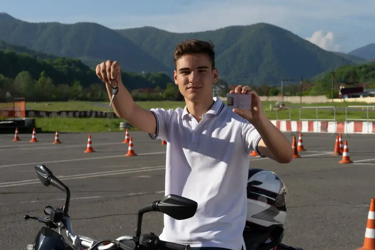Joven muestra carnet y llaves, listo para conducir moto de 125cc. El paisaje monta&ntilde;oso y conos de tr&aacute;fico sugieren una autoescuela.