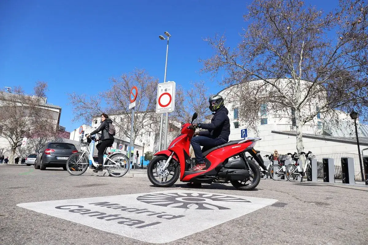 Un motorista en una moto roja espera en un cruce con se&ntilde;alizaci&oacute;n de Madrid Central. La normativa de aparcamiento de motos es visible en las se&ntilde;ales.