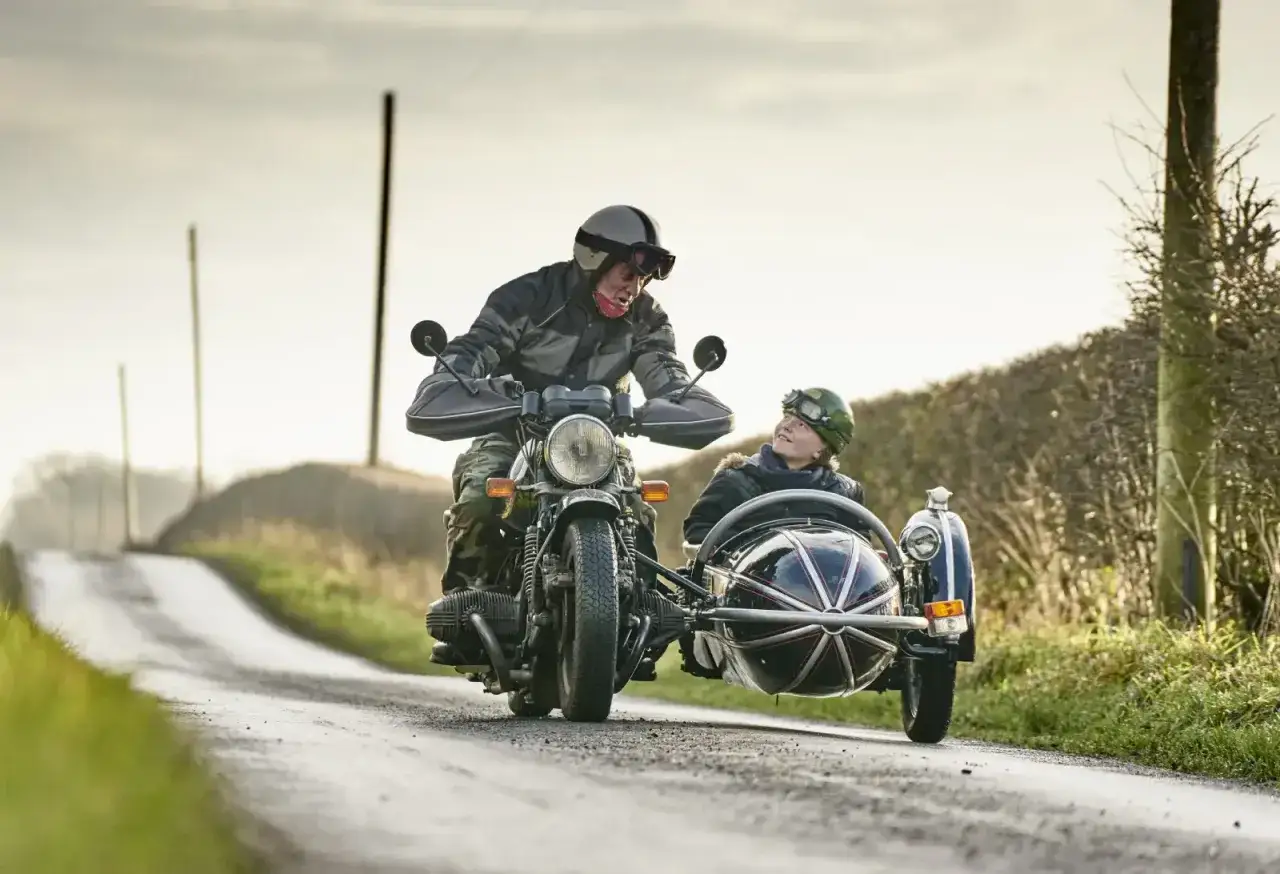 Un hombre y un niño disfrutan de un paseo en moto con sidecar por un camino rural. El niño mira al hombre con una sonrisa.
