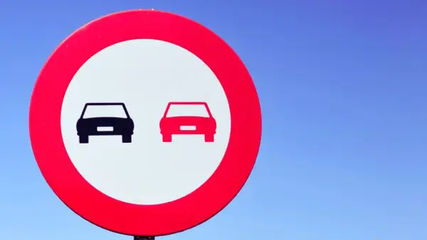 Señal de tráfico: dos coches, uno negro y otro rojo, en un círculo rojo sobre fondo blanco. Indica donde está prohibido adelantar.