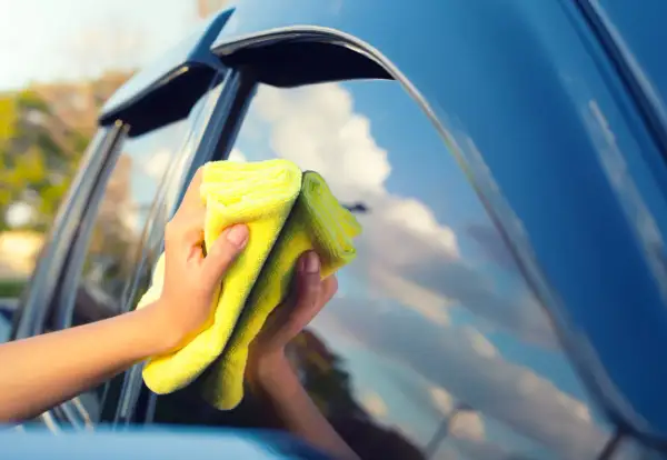 Manos limpiando los cristales del coche con un paño amarillo. El cielo azul y las nubes se reflejan en el cristal.