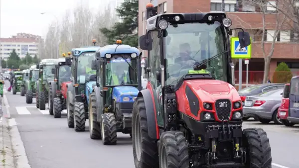 Una fila de tractores, algunos verdes y uno rojo al frente, circulan por una calle urbana. Parece que un tractor puede ir por autovía.