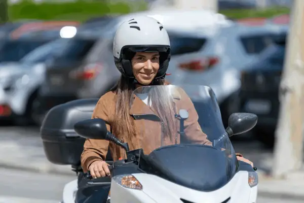 Mujer sonriente con casco blanco y chaqueta marrón en moto. Averigua el precio del carnet de moto A2.