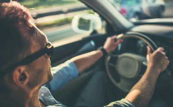 Hombre con gafas de sol conduce, notando que mi coche vibra a partir de 100 km/h. El sol ilumina su cabello.